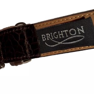 Może przedstawiać: Brązowy sk&oacute;rzany pasek z czarną etykietą, na kt&oacute;rej widnieje napis "BRIGHTON" srebrnymi literami. Etykieta ma srebrny wz&oacute;r ryby.