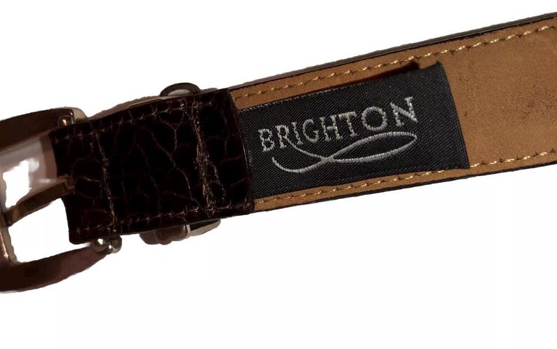 Może przedstawiać: Brązowy sk&oacute;rzany pasek z czarną etykietą, na kt&oacute;rej widnieje napis "BRIGHTON" srebrnymi literami. Etykieta ma srebrny wz&oacute;r ryby.
