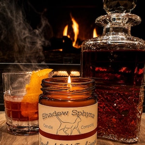 May include: A brown glass candle jar with a label reading Shadow Sphynx Candle Company Smoked Old Fashioned and ShadowSphynxCandles.etsy.com, featuring a cat illustration. Beside it sits a crystal decanter and a glass of amber liquid with orange peel.