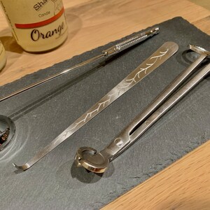 May include: A set of three silver candle care tools on a black slate surface. The tools include a wick trimmer, a wick dipper, and a pair of candle shears.
