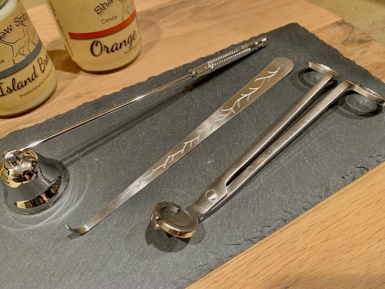 May include: A set of three silver candle care tools on a black slate surface. The tools include a wick trimmer, a wick dipper, and a pair of candle shears.