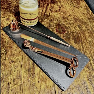 May include: A black slate tray with a gold candle snuffer, a gold wick trimmer, and a gold candle holder. The tray is on a wooden surface. The candle jar is labelled "Shadow Sphinx" and "Smoked Old Fashioned".