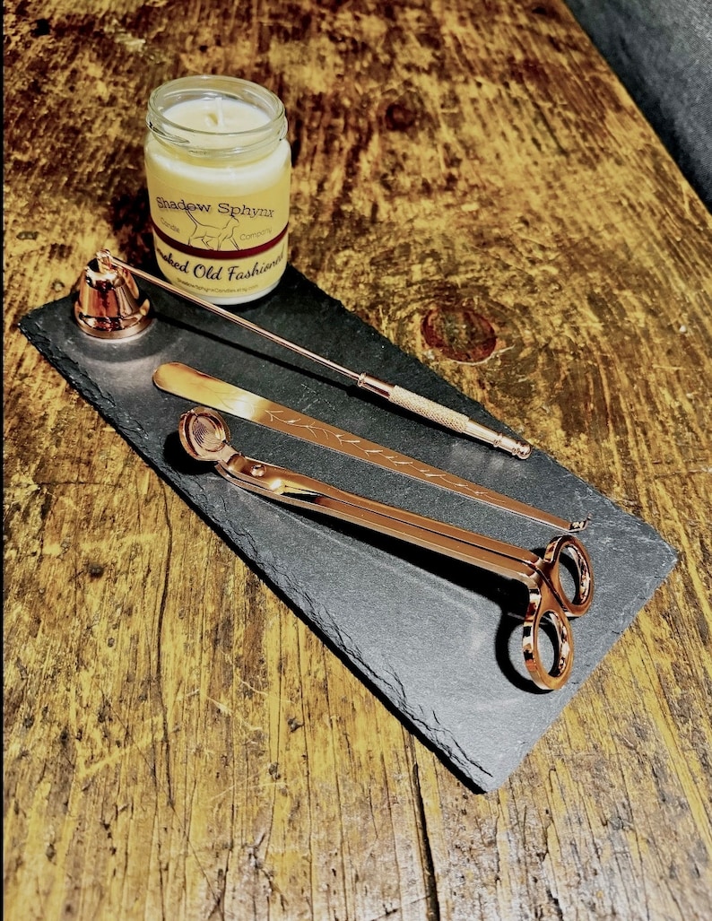 May include: A black slate tray with a gold candle snuffer, a gold wick trimmer, and a gold candle holder. The tray is on a wooden surface. The candle jar is labelled "Shadow Sphinx" and "Smoked Old Fashioned".