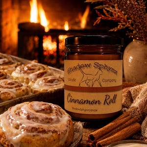 May include: A brown glass jar candle with a label reading Shadow Sphynx Candle Company Cinnamon Roll. The label features a line drawing of a cat. The candle is positioned near a plate with a glazed cinnamon roll and several cinnamon sticks.