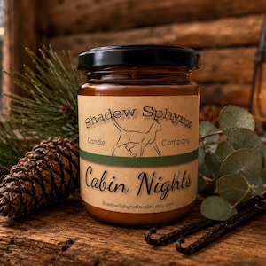 May include: A brown glass candle jar with a black lid and a tan label. The label features the text Shadow Sphynx Candle Company, a line drawing of a cat, and the text Cabin Nights. The jar is surrounded by a pinecone, pine needles, and vanilla beans.