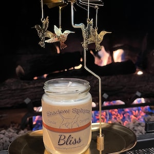 May include: A gold metal candle holder with a spinning design featuring hummingbirds. The candle holder is holding a glass jar candle with a white label that reads "Shadow Sphynx Candle Company" and "Bliss".