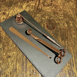 May include: A set of three gold-toned candle accessories on a black slate surface. The set includes a candle snuffer, a pair of candle wick trimmers, and a candle holder.