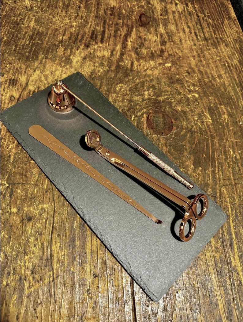 May include: A set of three gold-toned candle accessories on a black slate surface. The set includes a candle snuffer, a pair of candle wick trimmers, and a candle holder.