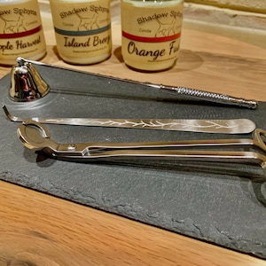 May include: A set of candle accessories on a black slate surface. The set includes a candle snuffer, a wick trimmer, and a pair of candle tongs. The candle snuffer is a bell-shaped device with a handle. The wick trimmer is a long, thin tool with a small blade at one end. The candle tongs are a pair of metal tongs with a spring-loaded mechanism.