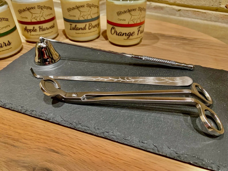 May include: A set of candle accessories on a black slate surface. The set includes a candle snuffer, a wick trimmer, and a pair of candle tongs. The candle snuffer is a bell-shaped device with a handle. The wick trimmer is a long, thin tool with a small blade at one end. The candle tongs are a pair of metal tongs with a spring-loaded mechanism.