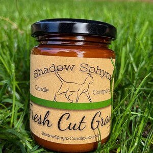 May include: A glass jar candle with a black lid and a tan label. The label features a line drawing of a cat, the text Shadow Sphynx Candle Company, and the text Fresh Cut Grass. The website ShadowSphynxCandles.etsy.com is printed at the bottom.