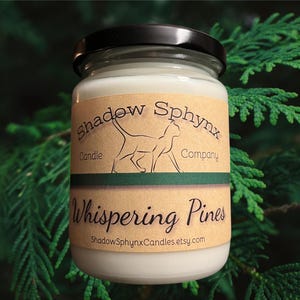 May include: A glass jar candle with a black lid, filled with white wax. The label reads "Shadow Sphynx Candle Company" with a cat silhouette and "Whispering Pines". The background is green foliage.