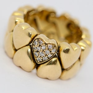 May include: A gold heart-shaped ring with a pave diamond heart in the center. The ring is made up of multiple connected heart shapes.