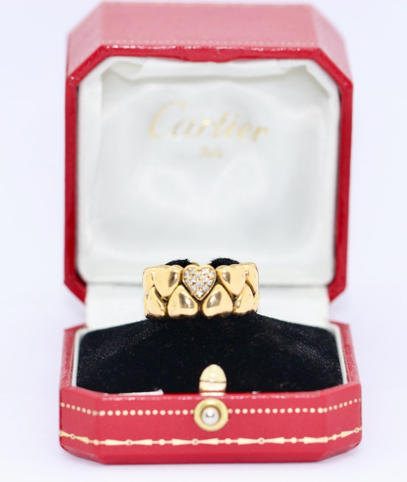 May include: A gold ring with a heart-shaped center set with diamonds. The ring is in a red Cartier box with a white interior.