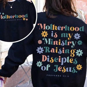 May include: Black sweatshirt with the text "Motherhood is my Ministry Raising Disciples of Jesus" in colorful letters and flower graphics. The text "Proverbs 22:6" is at the bottom. A smaller image shows the text "Motherhood is Kingdom Work".