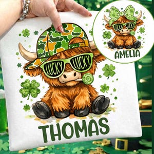 Op de afbeelding: Wit kussen met een cartoon Highland-koe met een groene zonnebril met "LUCKY" en een camouflage hoed. De koe houdt een groene lolly vast. De naam "THOMAS" staat in het groen onderaan. Een kleinere afbeelding van een koe met de naam "AMELIA" staat rechtsboven.