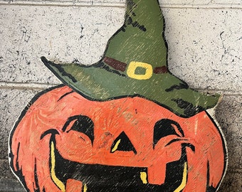 Puede incluir: Una calabaza de madera con una cara pintada de naranja, ojos negros y una amplia sonrisa con dientes. La calabaza lleva un sombrero de bruja verde con una banda marrón y una hebilla amarilla.