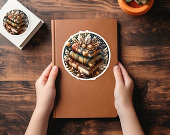 May include: A sticker depicting a stack of books with gold detailing and a floral wreath design. The sticker is placed on a brown notebook, with a matching sticker on a white square. A small potted plant sits nearby on a wooden surface.