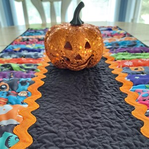 May include: A glittery orange jack-o'-lantern pumpkin with a black stem sits on a quilted table runner. The runner features a central black panel with stippled stitching, flanked by colorful Halloween-themed fabric and orange rickrack trim.