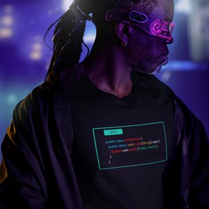 Pode incluir: Uma pessoa vestindo uma camiseta preta com um trecho de código impresso nela. O código está escrito na linguagem de programação Java e exibe o clássico programa "Hello, World!"