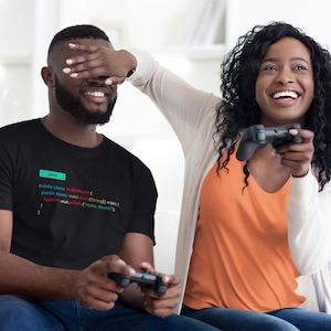 May include: A couple playing video games on a couch. The man is wearing a black t-shirt with code printed on it. The woman is wearing an orange shirt and a white cardigan. They are both smiling and laughing.