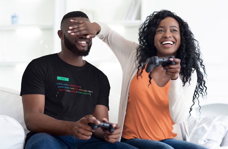 May include: A couple playing video games on a couch. The man is wearing a black t-shirt with code printed on it. The woman is wearing an orange shirt and a white cardigan. They are both smiling and laughing.