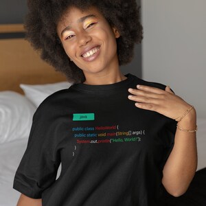 Puede incluir: Una persona lleva una camiseta negra con el código de un programa "Hello World" impreso en ella. El código está escrito en Java e incluye el texto "public class HelloWorld", "public static void main", "String[] args", y "System.out.println("Hello, World!");".