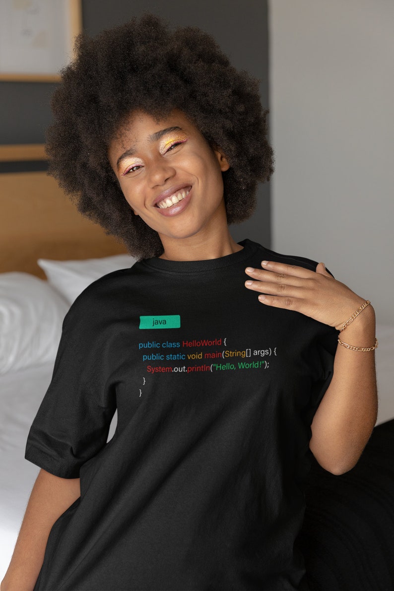 May include: A person wearing a black t-shirt with the code for a "Hello World" program printed on it. The code is written in Java and includes the text "public class HelloWorld", "public static void main", "String[] args", and "System.out.println("Hello, World!");".