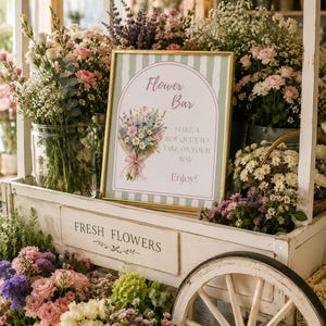 May include: A gold-framed sign with a green and white striped background features an illustration of a floral bouquet and the text Flower Bar, MAKE A BOUQUET TO TAKE ON YOUR WAY, and Enjoy! The sign sits on a white wooden cart with wheels.