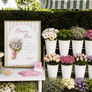 May include: A gold-framed sign with a green striped background and an arched illustration of a flower bouquet, featuring the text Flower Bar, Make a bouquet to take on your way, Enjoy. Beside it are white buckets filled with assorted fresh flowers.