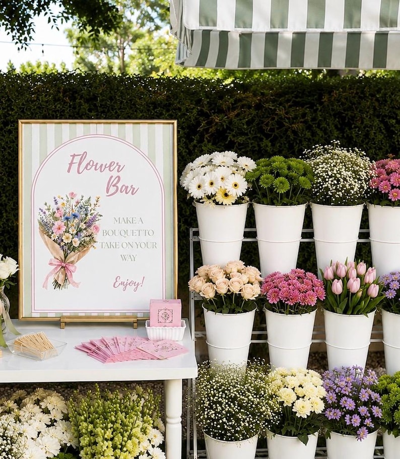 May include: A gold-framed sign with a green striped background and an arched illustration of a flower bouquet, featuring the text Flower Bar, Make a bouquet to take on your way, Enjoy. Beside it are white buckets filled with assorted fresh flowers.