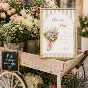May include: A rectangular sign in a gold frame with a green and white striped background, featuring an illustration of a flower bouquet and the text: Flower Bar, MAKE A BOUQUET TO TAKE ON YOUR WAY, Enjoy! The sign sits on a white wooden cart.