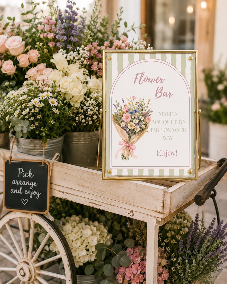 May include: A rectangular sign in a gold frame with a green and white striped background, featuring an illustration of a flower bouquet and the text: Flower Bar, MAKE A BOUQUET TO TAKE ON YOUR WAY, Enjoy! The sign sits on a white wooden cart.