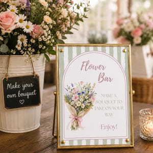 May include: A gold-framed sign with a green and white striped background featuring an illustration of a wildflower bouquet and the text: Flower Bar, MAKE A BOUQUET TO TAKE ON YOUR WAY, Enjoy!. The sign is positioned next to a white bucket of flowers.