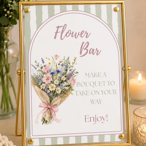 May include: A rectangular sign in a gold metal frame featuring a green and white striped background. The sign displays the text "Flower Bar," "MAKE A BOUQUET TO TAKE ON YOUR WAY," and "Enjoy!" alongside a colourful illustration of a flower bouquet.
