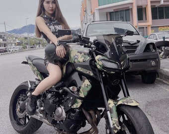 Może przedstawiać: Kobieta siedzi na czarno-zielonym motocyklu w kamuflażu. Motocykl ma czarną szybę i czarne siedzenie. Kobieta ma na sobie czarną koszulę na ramiączkach z białym napisem i czarne spodenki.