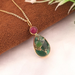 May include: A gold necklace with a pendant. The pendant has a teardrop-shaped green stone with gold detailing and a small, round, red stone. The necklace is shown on a brown surface.