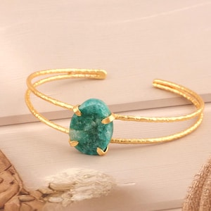 May include: Gold-coloured cuff bracelet with a large, oval teal-green gemstone. The bracelet has a hammered texture and a double-band design. The gemstone is held in place by four gold prongs.