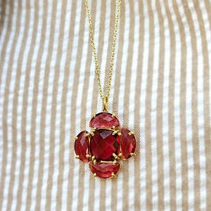 May include: A gold-colored necklace with a pendant featuring a cluster of red gemstones. The pendant has a central, square-cut stone surrounded by four oval-shaped stones. The necklace chain is delicate and hangs against a striped background.