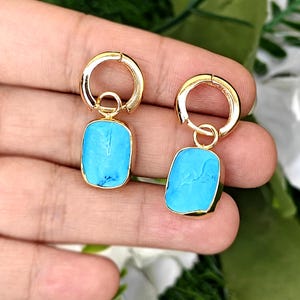 Puede incluir: Un par de pendientes de aro dorados con colgantes rectangulares turquesa. Los pendientes tienen un diseño sencillo, con un pequeño aro que se conecta a una piedra rectangular. Las piedras turquesas tienen un aspecto ligeramente texturizado.