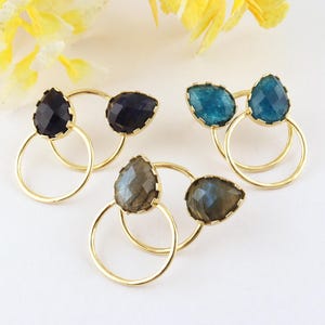 Pode incluir: Três pares de brincos dourados, cada um com dois aros circulares entrelaçados e uma pedra preciosa facetada em forma de pera. As pedras são azul-escuras, azul-petróleo e cinza iridescente, montadas em armações de metal dourado polido.