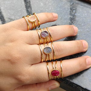Puede incluir: Cuatro anillos dorados en los dedos, cada uno con un diseño de banda fina de múltiples hilos. Los anillos están engastados con piedras preciosas ovaladas de textura rugosa en varios colores, incluyendo una piedra translúcida, una morada, una gris y una roja.