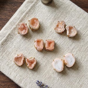 May include: Five pairs of stud earrings featuring irregular, peach-colored druzy stones set in gold-toned metal prongs. The stones have a crystalline, textured center and smooth, rounded edges, arranged in a cluster on a light-colored fabric surface.