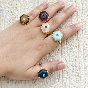 May include: A hand displaying six gold-toned rings. Each ring features a unique, hexagonal gemstone in shades of brown, purple, pink, blue, and teal. Some rings have a small, clear stone in the center. The rings are set against a neutral background.