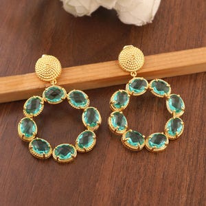 May include: A pair of gold-coloured hoop earrings. Each earring has a textured, circular stud and a hoop design decorated with oval, teal-coloured gemstones. The earrings are displayed on a wooden surface.