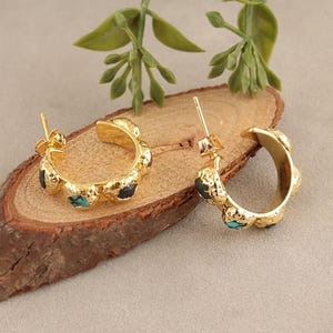 Peut inclure: Une paire de créoles dorées présentant une surface texturée et organique. Chaque créole est incrustée de petits morceaux irréguliers de turquoise bleue et de pierres sombres. Les boucles d'oreilles incluent des fermoirs à tige classiques.