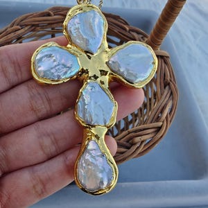 May include: A cross-shaped pendant necklace featuring four irregular, iridescent white baroque pearls set in a polished gold-tone metal frame. The pendant hangs from a delicate gold-tone chain, showcasing a textured, organic design with a lustrous finish.