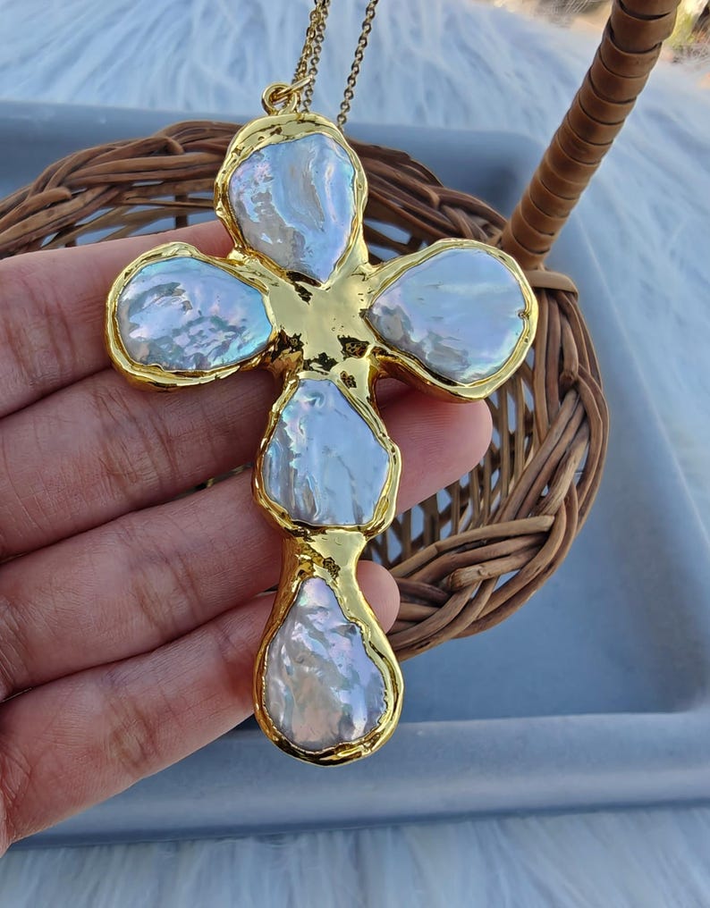 May include: A cross-shaped pendant necklace featuring four irregular, iridescent white baroque pearls set in a polished gold-tone metal frame. The pendant hangs from a delicate gold-tone chain, showcasing a textured, organic design with a lustrous finish.