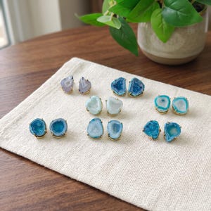 May include: A collection of six pairs of stud earrings featuring irregular, geode-style agate slices in various shades of blue, teal, and white. Each stone is encased in a gold-toned metal setting with small prongs securing the edges.