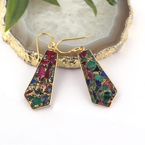 Puede incluir: Un par de pendientes dorados con una piedra rectangular en forma de trapecio. La piedra es un mosaico de colores rojo, verde, azul y dorado. Los pendientes tienen un cierre de gancho.